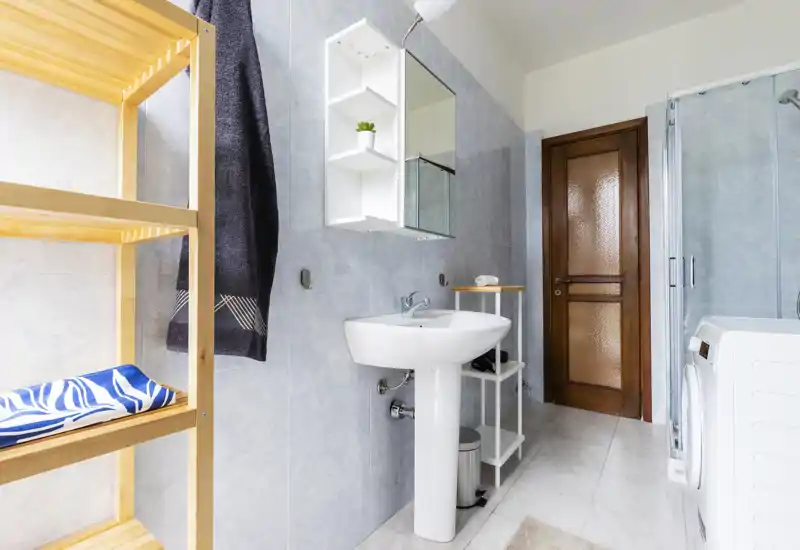 Bright, modern bathroom with a pedestal sink, mirrored cabinet, shower enclosure and washing machine; light tiles and wooden door create a clean, functional space.