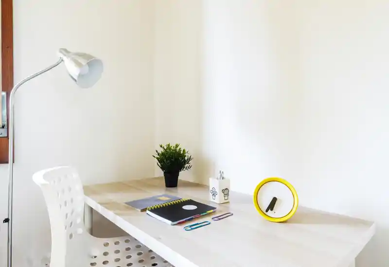 Compact, bright workspace with a light wood desk, white perforated chair, a small plant, stationery and a yellow-accented clock — ideal for focused studying or remote work.