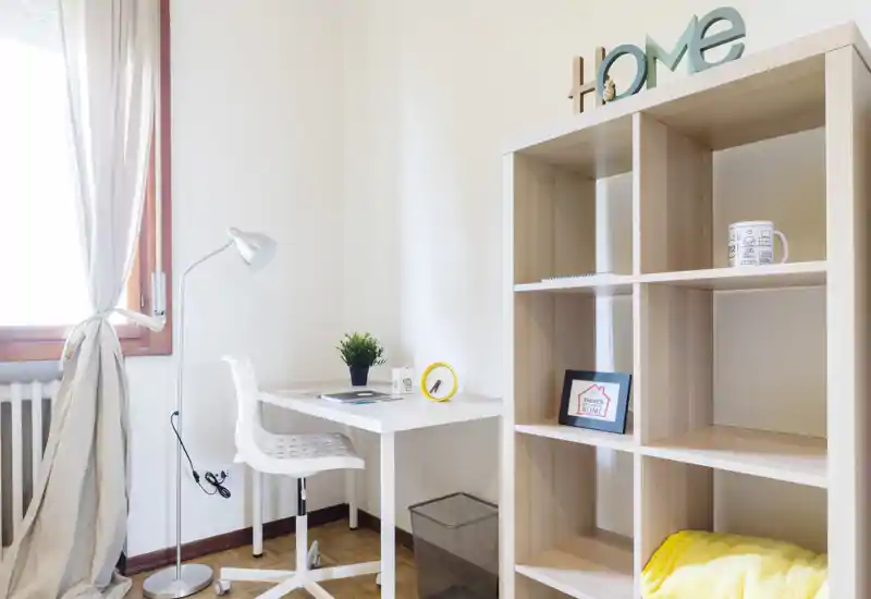 Bright, minimal living area with a compact desk and shelving unit — ideal for a home office corner. Natural light from the window and simple décor create a fresh, functional space.