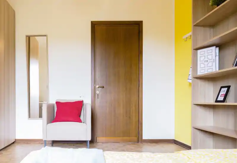 Bright, tidy bedroom corner with a wooden door, an armchair with a red cushion and a partial view of the bed in the foreground; warm wood shelving and a yellow accent wall add character.