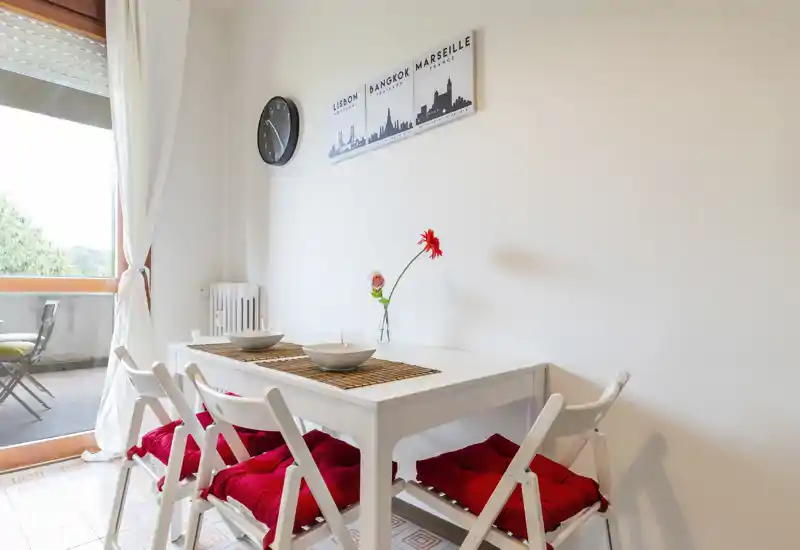 Bright, cozy dining corner with a white table, four folding chairs with red cushions, simple table settings and a vase with a single flower; large window/door opens to a small balcony.