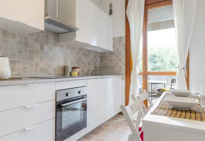 Bright, modern kitchen with white cabinetry, built-in oven and a small dining table by a balcony — ideal for cozy meals with natural light.
