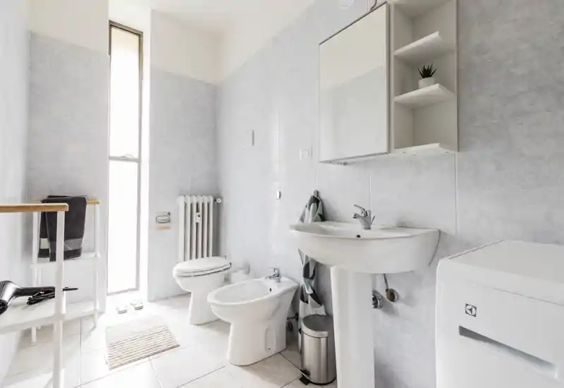 Bright, modern bathroom with a pedestal sink, bidet and toilet next to a tall window. Clean tiles and minimal shelving create a fresh, airy feel ideal for rental listings.