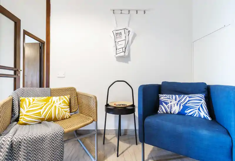 Bright, stylish living corner with a blue armchair and woven chair separated by a small black side table; decorative cushions and a throw create a cozy, modern sitting area.