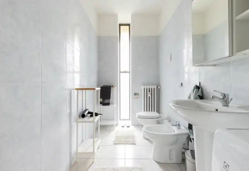 Bright, modern bathroom with white fixtures including sink, bidet and toilet, pale tiled walls and a tall narrow window providing natural light.