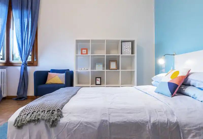 Bright, modern bedroom with a comfortable double bed, colorful throw pillows and a cozy armchair by the window. Clean lines, soft textiles and a light-blue accent wall create a fresh, welcoming atmosphere ideal for rental listings.