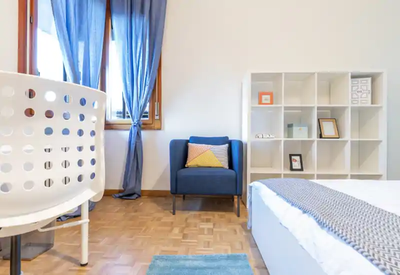 Bright, minimal bedroom with a comfortable bed dressed in a knit throw, a navy armchair and a white cubed shelving unit — ideal for a calm, modern stay.