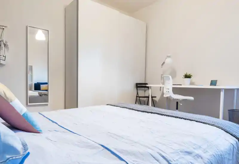 Bright, modern bedroom with a large bed in the foreground, simple white wardrobe and a neat study desk with chair and lamp — clean, airy and well-styled for rental photos.