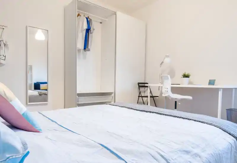 Bright, modern bedroom with a large bed in the foreground, minimalist wardrobe, and a neat study desk. Soft textiles and colorful cushions give a welcoming, contemporary feel.