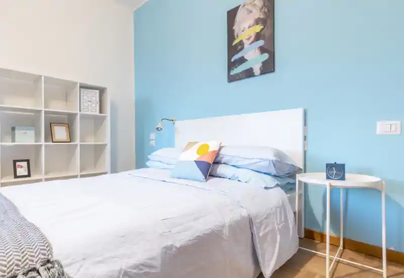 Bright, modern bedroom with a neatly made double bed against a soft blue accent wall, decorative cushion, bedside table with a clock and built-in shelving for storage — ideal for a cozy rental listing.