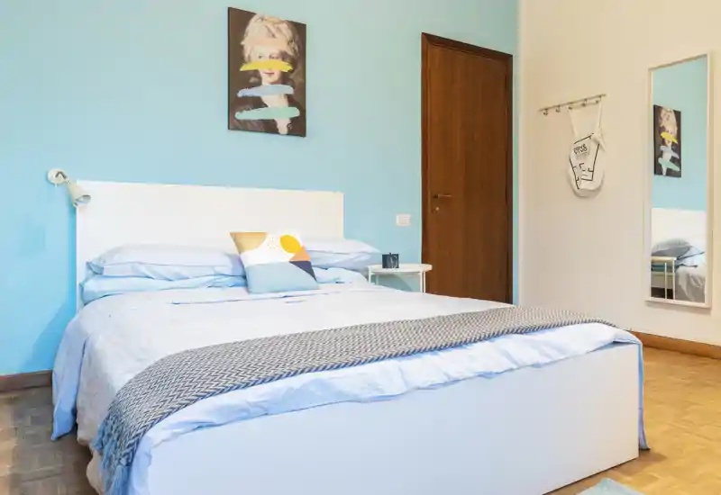 Bright, modern bedroom with a neatly made double bed, pastel blue accent wall and minimalist decor. The room features a bedside table, wall lamp and a full-length mirror that reflects part of the space.