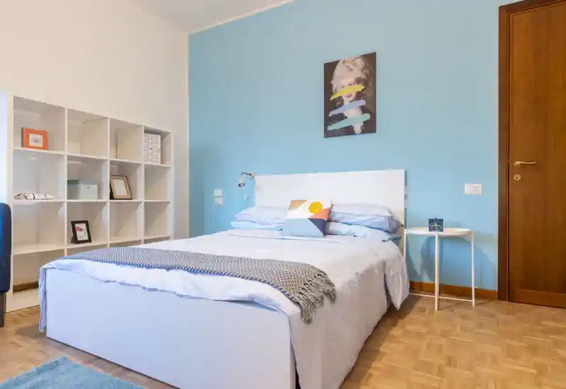 Bright, modern bedroom with a neatly made double bed against a soft blue accent wall, simple white headboard, bedside table and shelving unit. Calm styling and natural light create a welcoming space ideal for rental photos.