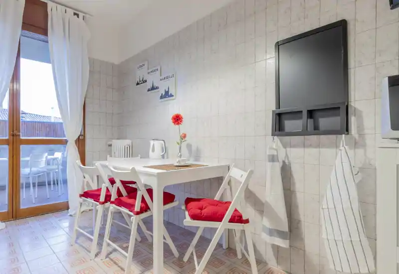 Bright, neatly staged dining area with a white table and four chairs dressed with red cushions; sliding door opens to a small terrace and natural light fills the space.