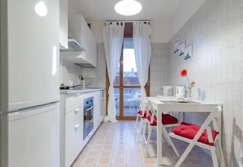 Bright, modern galley kitchen with white cabinets, built-in oven, fridge and a small dining table with red-cushioned chairs opening onto a balcony.