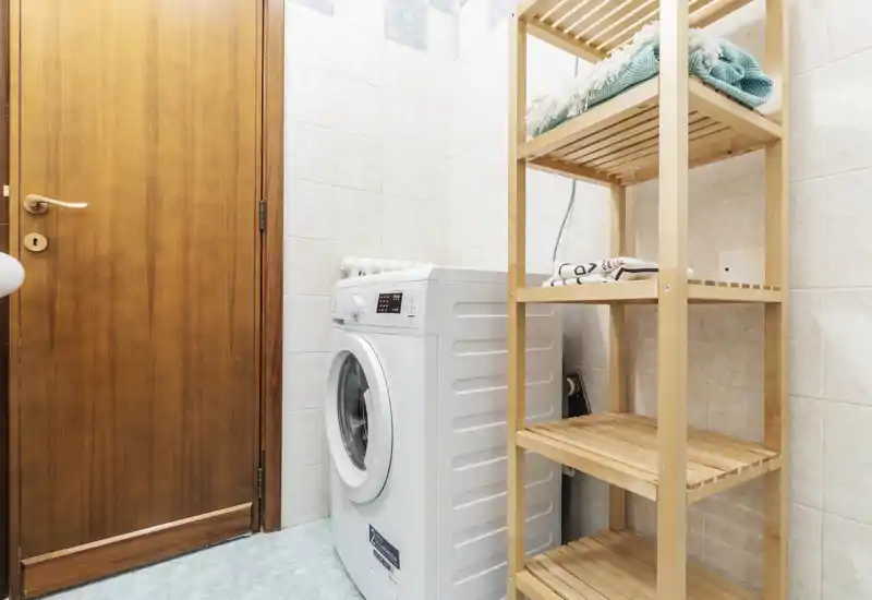 Compact laundry/utility corner with a front-loading washing machine and wooden shelving for towels and supplies.