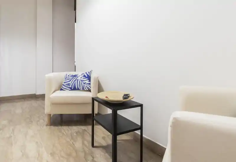 Small, bright sitting area with two cream armchairs, a decorative blue cushion and a compact black side table — ideal for a cozy corner or waiting area.