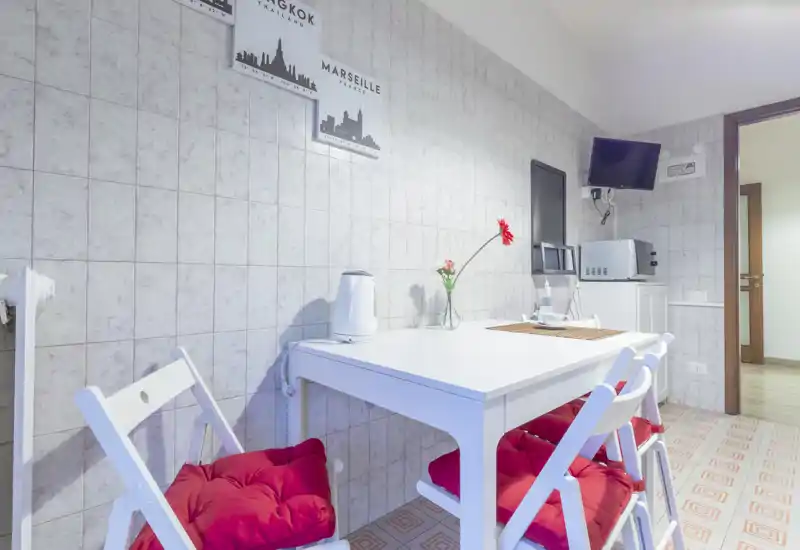 Small, bright kitchen dining corner with a white table, foldable chairs topped with red cushions, microwave and kettle — a neat, cozy space ideal for casual meals.