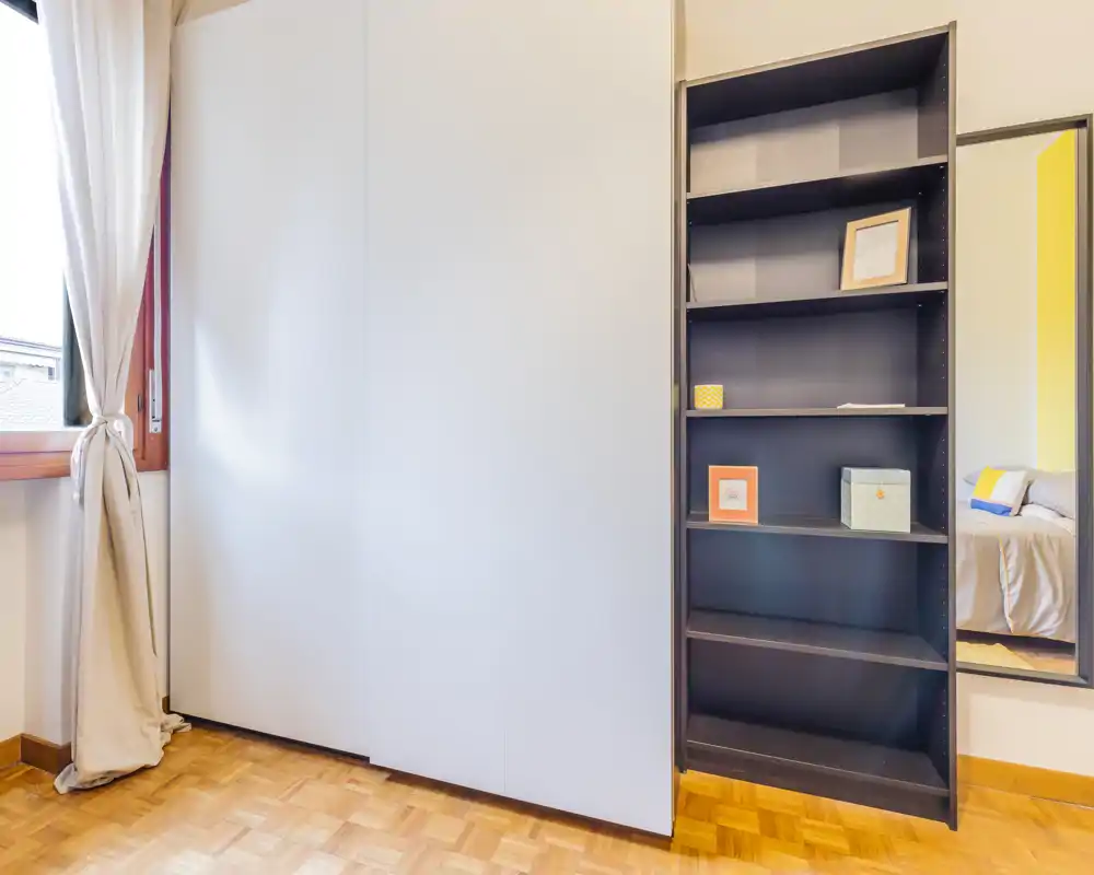 Bright bedroom corner with a large built-in wardrobe, shelving and a mirror that reflects part of the bed; natural light from the window creates a clean, airy feeling.