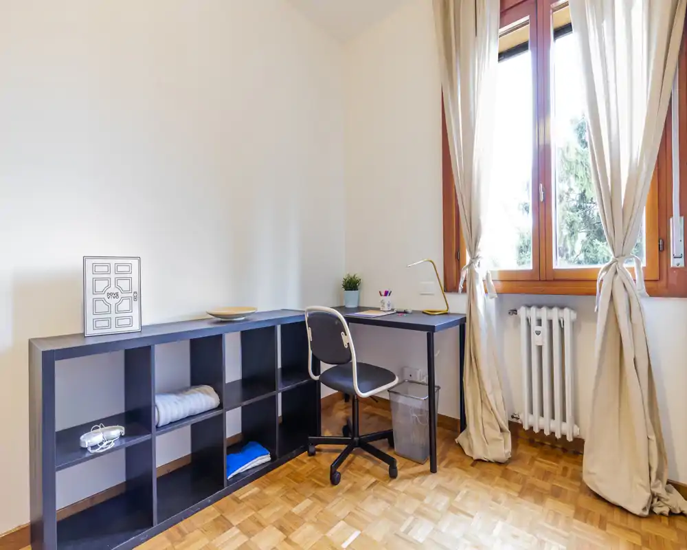 Bright, minimal living/working corner with a desk, shelving unit and a large curtained window letting in natural light.