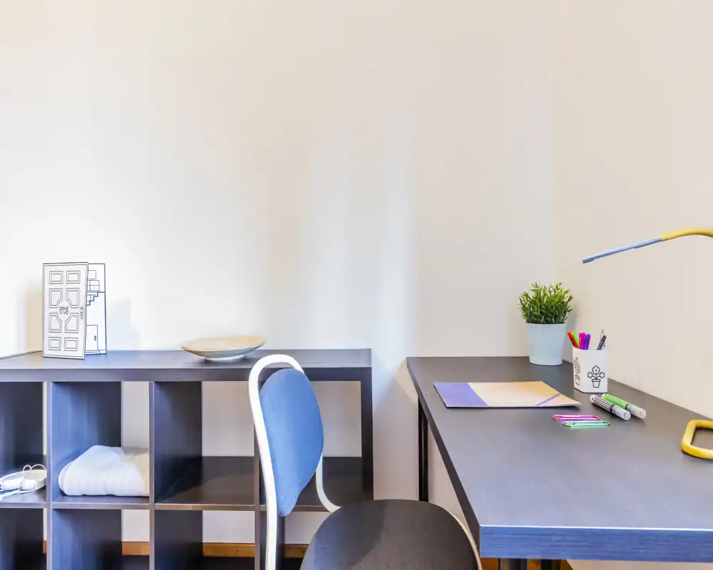 Bright, tidy study corner with a dark desk, chair, desk lamp and shelving. Small plant and stationery add a lived-in, functional touch.