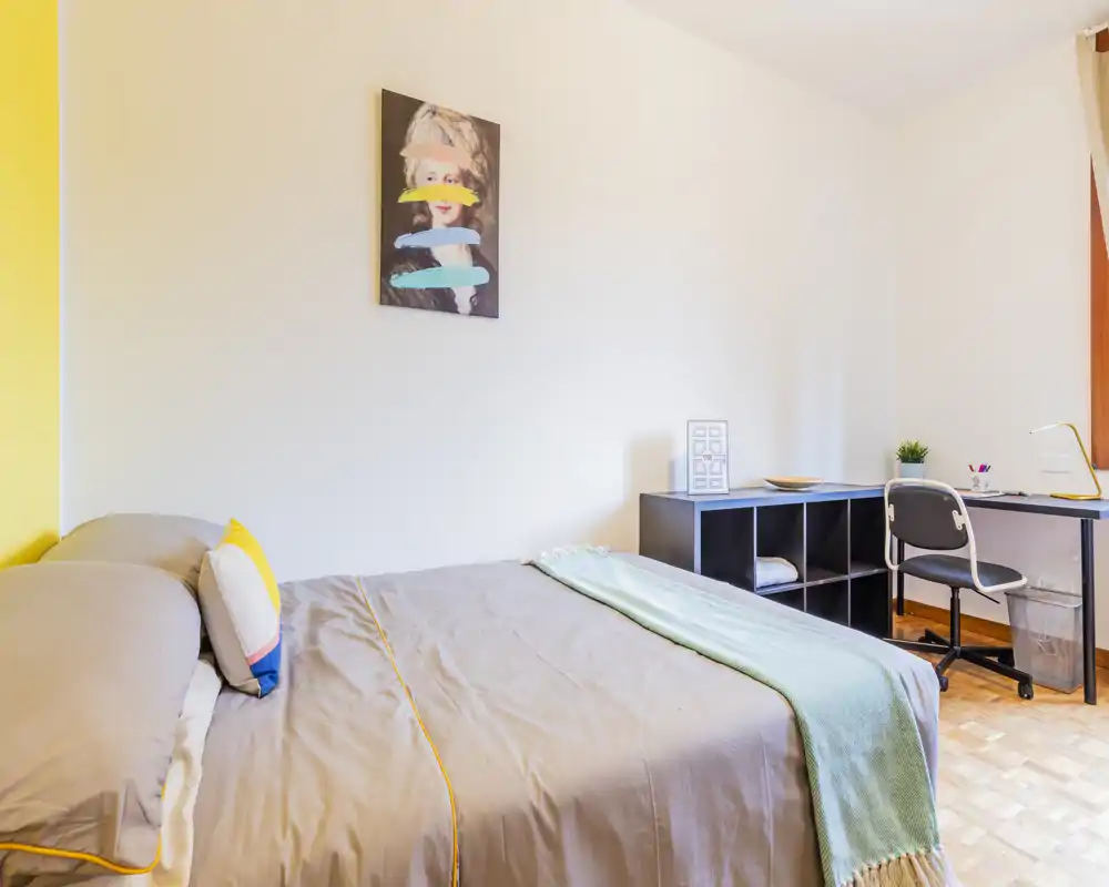 Bright, modern bedroom with a neatly made double bed, colorful cushions and a soft throw; workspace with desk and shelving visible in the corner.