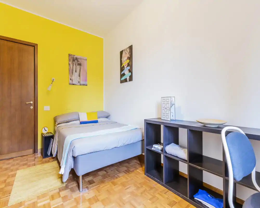 Bright, modern bedroom with a neatly made double bed against a cheerful yellow accent wall, parquet flooring and a practical shelving desk area.