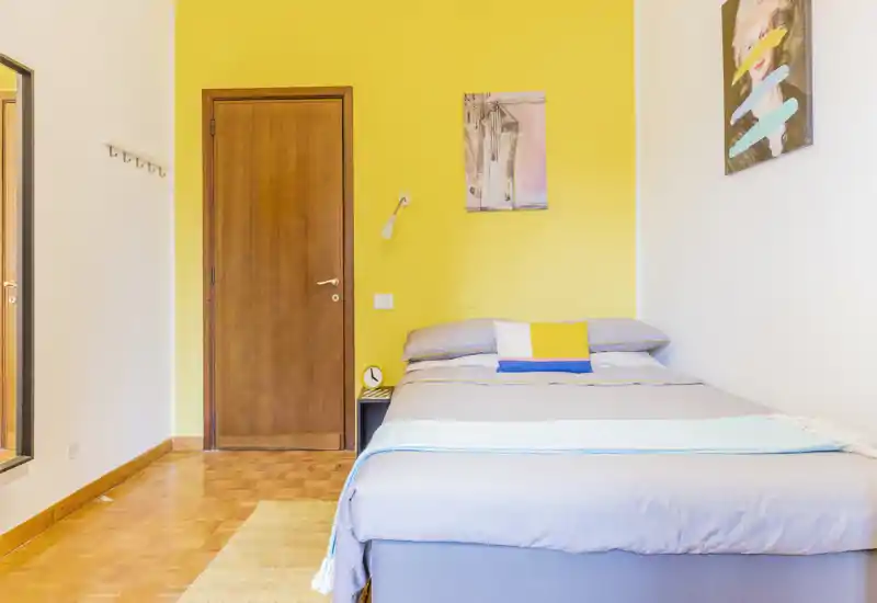 Bright, modern bedroom with a neatly made double bed against a vibrant yellow accent wall. Soft textiles, wall art and bedside lamp create a cozy, well-styled atmosphere ideal for rental photos.
