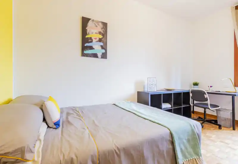 Bright, modern bedroom with a neatly made double bed, colorful cushions and a soft throw; workspace with desk and shelving visible in the corner.