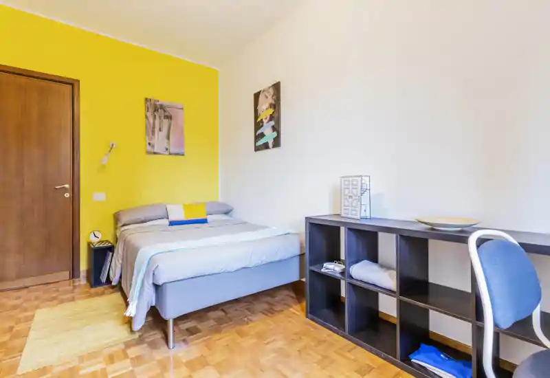 Bright, modern bedroom with a neatly made double bed against a cheerful yellow accent wall, parquet flooring and a practical shelving desk area.
