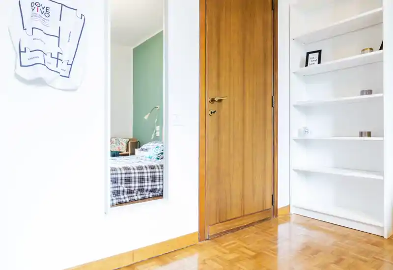 Bright, tidy bedroom view seen through an interior opening: you can spot a patterned bedspread, bedside lamp and cushions framed by a white doorway and warm wood door.