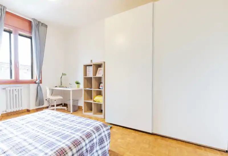 Camera da letto luminosa e ordinata con letto matrimoniale in primo piano, una scrivania vicino alla finestra e ampi armadi a muro. Parquet pulito e luce naturale la rendono perfetta per un annuncio in affitto.