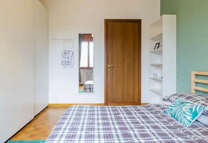 Bright, well-styled bedroom with a large plaid bed in the foreground, wooden door and wardrobe, plus tasteful decorative pillows and shelving.