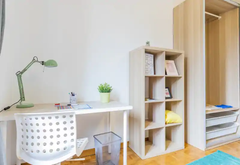 Compact bedroom corner with a white study desk, perforated chair, open shelving unit and an open wardrobe. Bright, minimal styling with a small plant and desk lamp ideal for a student or home office corner in a bedroom.