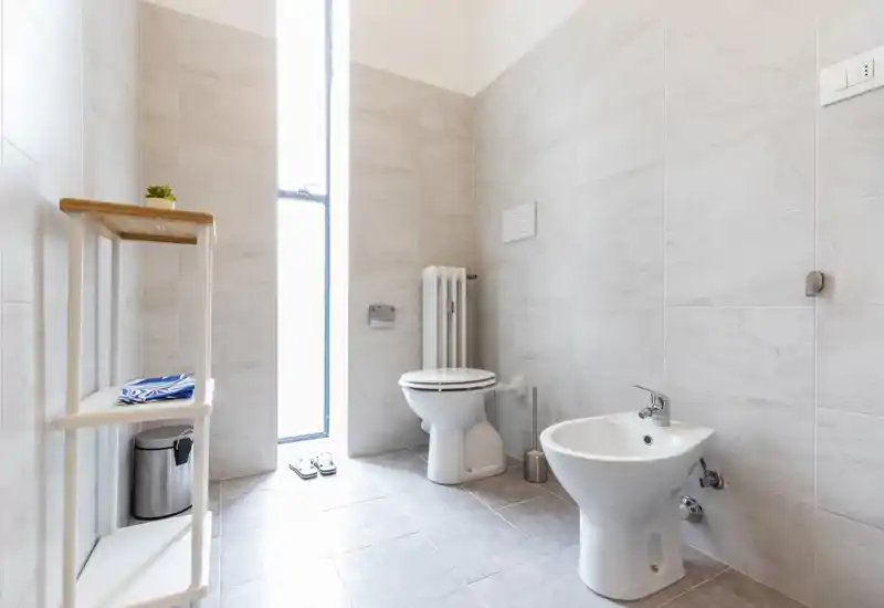 Bright, modern bathroom with a toilet and bidet, light tile walls and floor, and a tall narrow frosted window providing natural light.