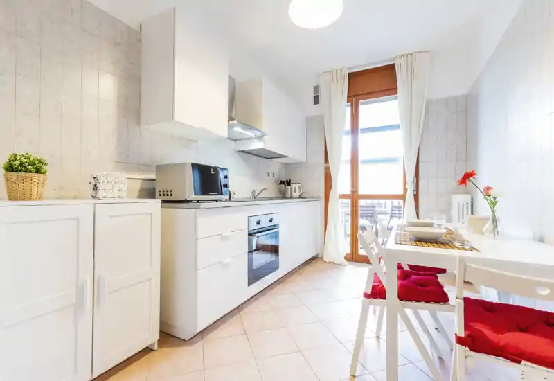 Bright modern kitchen with white cabinets, built-in oven and a small dining area by a glass door that opens to a balcony. Clean, well-lit space with cozy red seat cushions and simple decor.