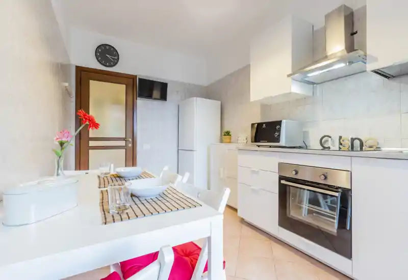 Bright, modern kitchen with white cabinets, oven, fridge and a small dining table set for two with decorative flowers — tidy and inviting for guests.