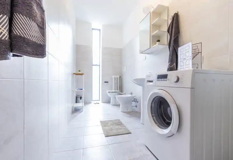 Bright modern bathroom with a front-loading washing machine, toilet, bidet and sink; light tiles and a tall narrow window create an airy, clean atmosphere.