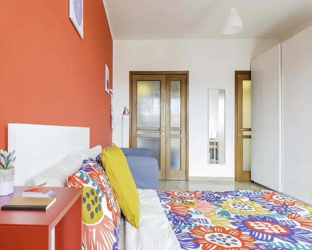 Camera da letto luminosa e colorata con copriletto decorato, cuscini e parete rossa d'accento; arredi moderni e molta luce naturale.