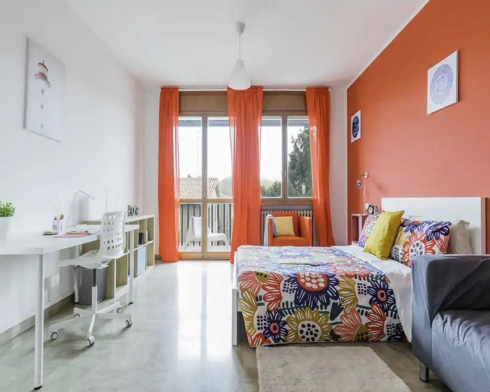 Camera da letto luminosa e vivace con letto matrimoniale e copriletto floreale, ampie finestre con tende rosse e una scrivania: moderna e accogliente.