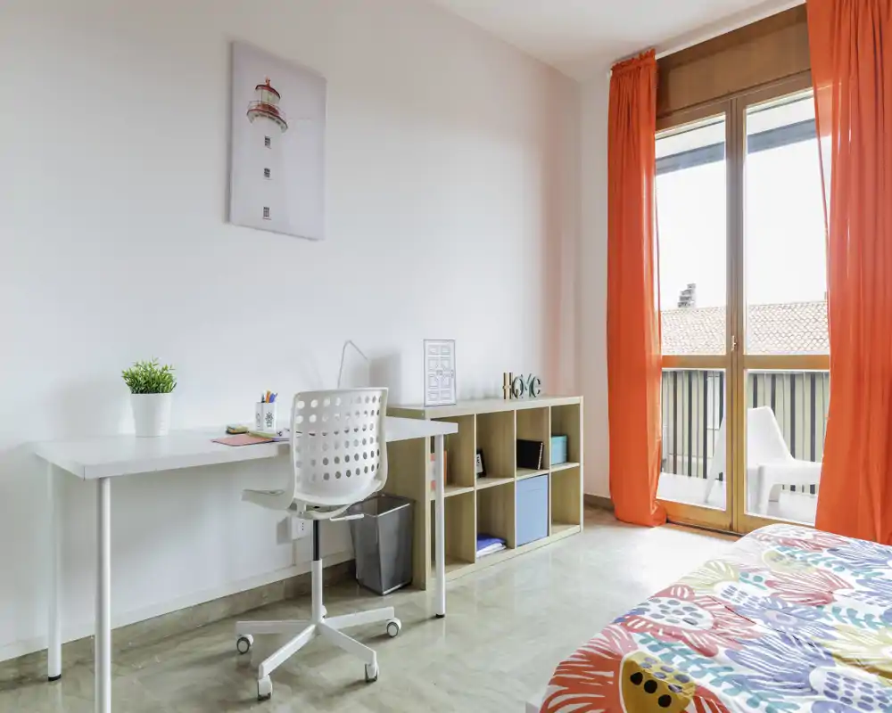Angolo camera luminoso e moderno con scrivania e accesso al balcone. Biancheria colorata e scaffalatura ordinata creano un ambiente fresco e accogliente, perfetto per un annuncio.