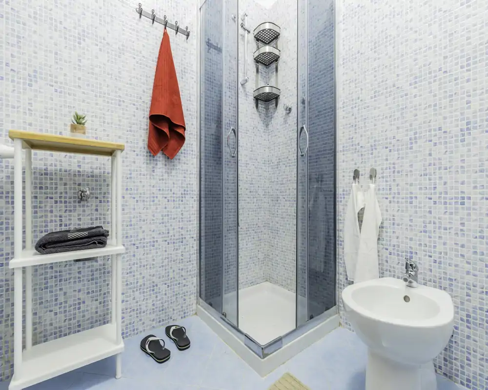 Bagno moderno e luminoso con box doccia angolare in vetro, bidet, pareti in mosaico e accessori ordinati per un soggiorno fresco e pulito.