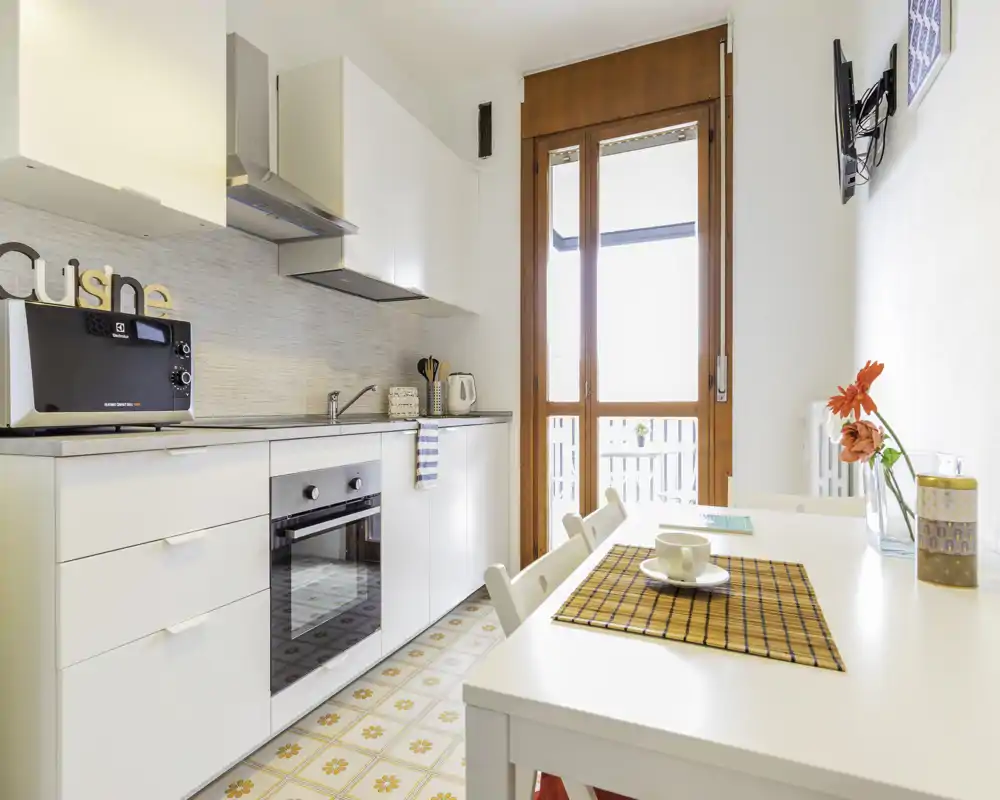 Cucina moderna e luminosa con forno integrato, microonde e un piccolo tavolo da pranzo vicino a una grande porta finestra che lascia entrare luce naturale. Pensili bianchi, pavimento a piastrelle decorate e dettagli semplici rendono l'ambiente accogliente per cucinare e consumare pasti informali.