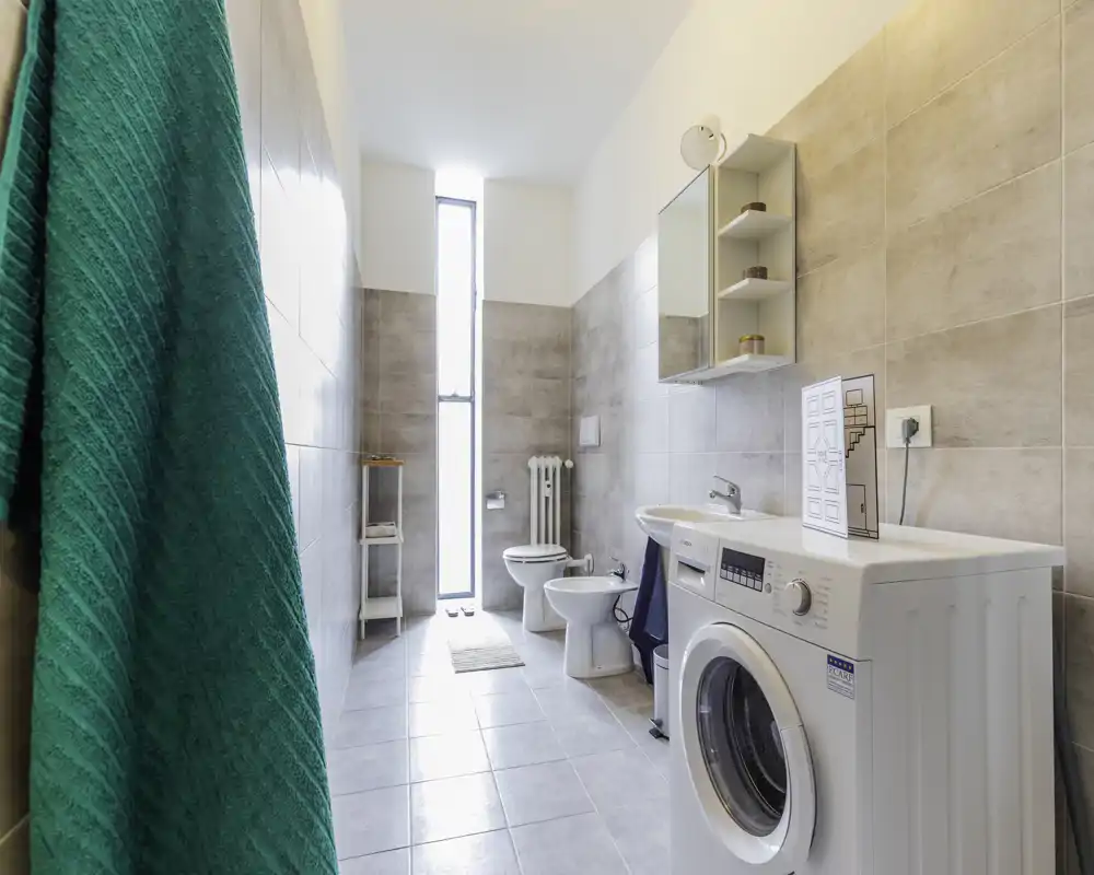 Bagno moderno e luminoso con lavatrice, wc, bidet e mobile pensile; una finestra stretta porta luce naturale e le piastrelle donano un aspetto pulito e contemporaneo.