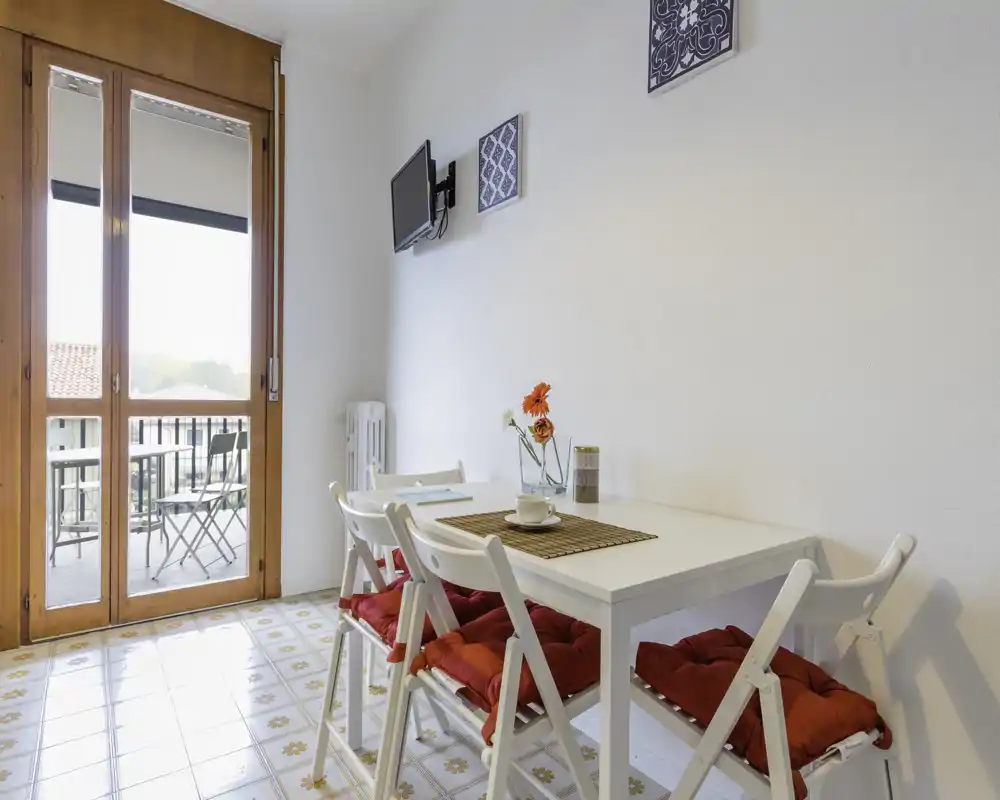 Angolo pranzo luminoso con tavolo bianco e quattro sedie con cuscini rossi, una mise en place semplice e un balcone che garantisce luce naturale.