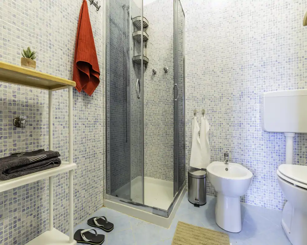 Bagno pulito e compatto con box doccia in vetro, bidet e WC, piastrelle a mosaico azzurro chiaro e accessori pratici.