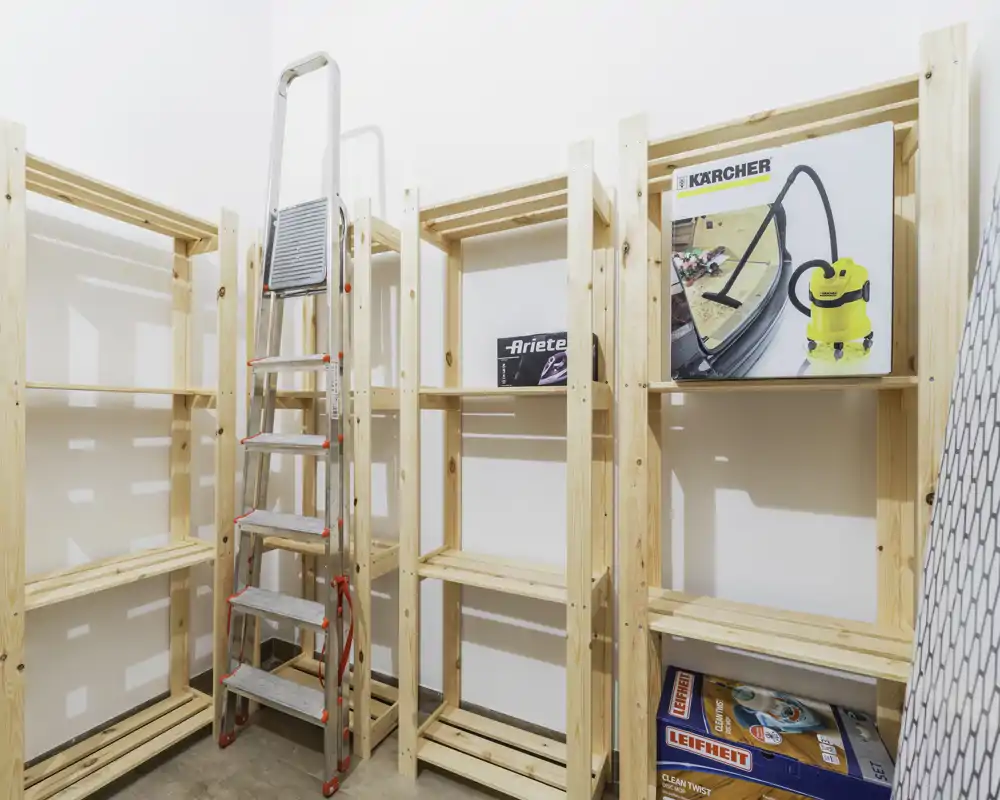 Piccolo locale di deposito/servizio con scaffalature in legno, una scala pieghevole e scatole con elettrodomestici; spazio luminoso e ordinato ideale per dispensa o detergenti.