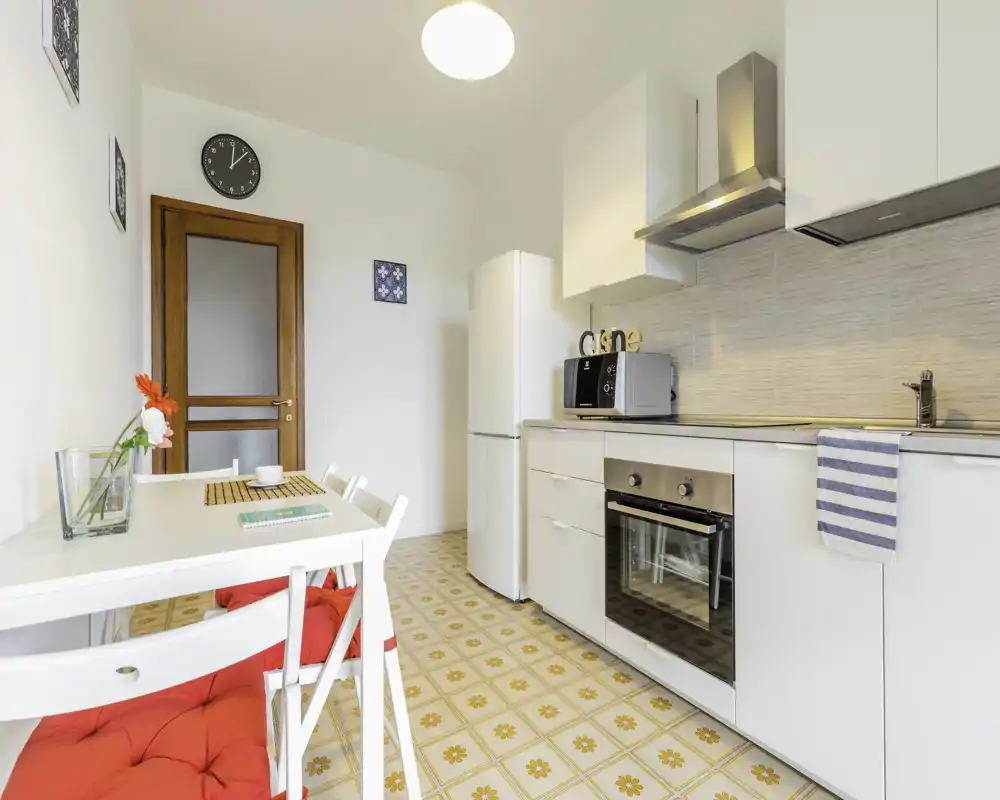 Cucina moderna e luminosa con mobili bianchi, forno integrato, frigorifero e un piccolo tavolo da pranzo con cuscini rossi — ambiente pulito e ben illuminato per pasti informali.