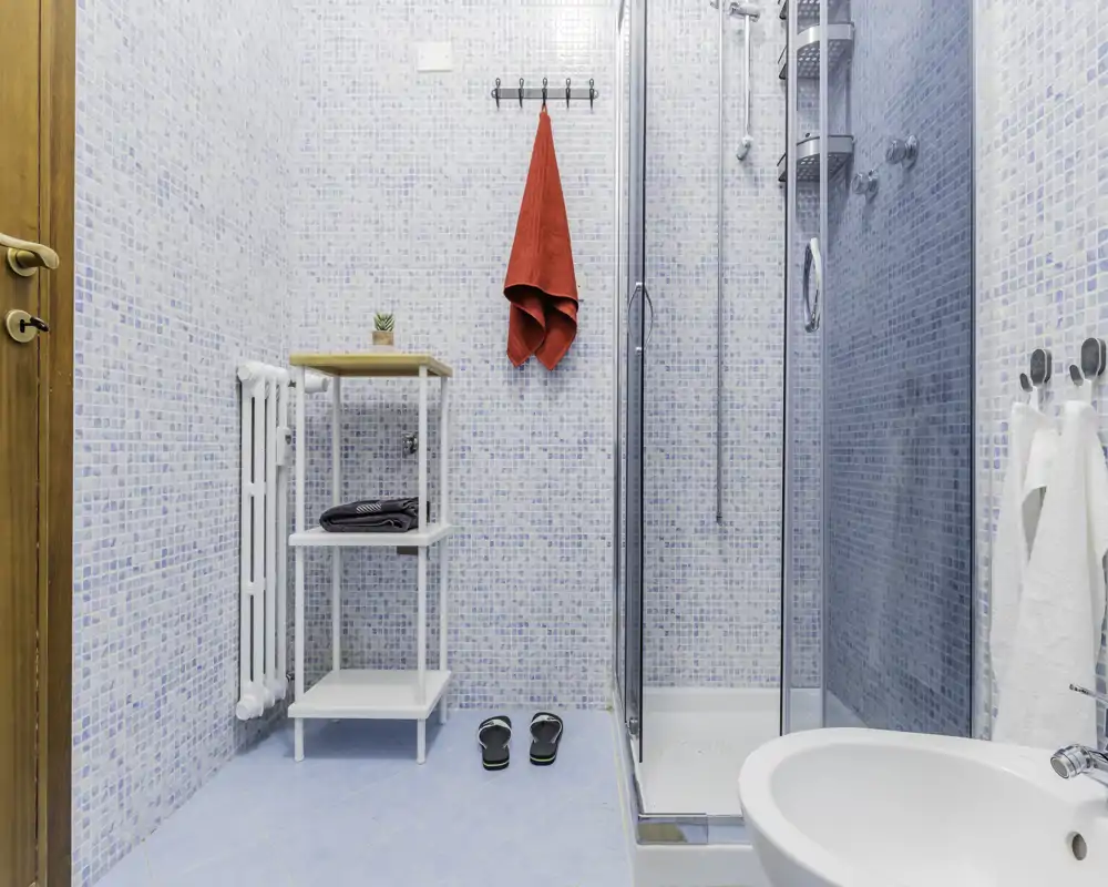 Bagno luminoso e moderno con box doccia in vetro, lavabo e piastrelle a mosaico azzurro. Mensole ordinate, ganci con asciugamani e un piccolo termosifone completano la disposizione funzionale.