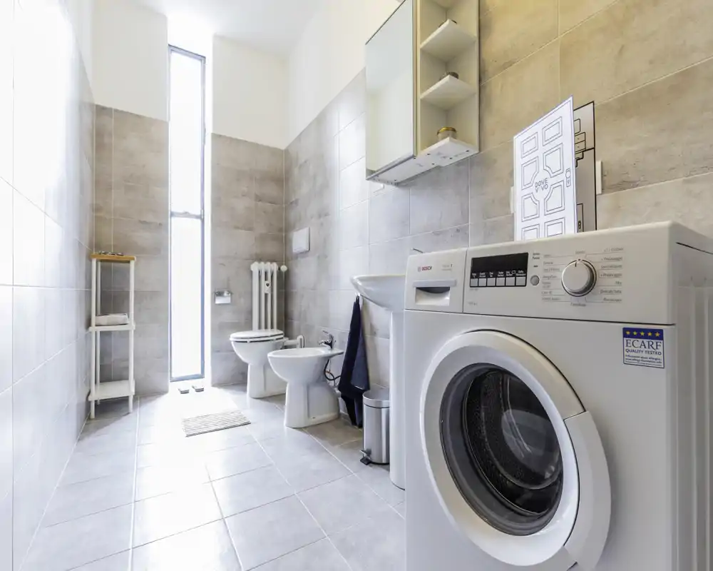 Bagno moderno e luminoso con piastrelle, WC, bidet, lavabo con pensile-specchio e una lavatrice a carica frontale in primo piano.