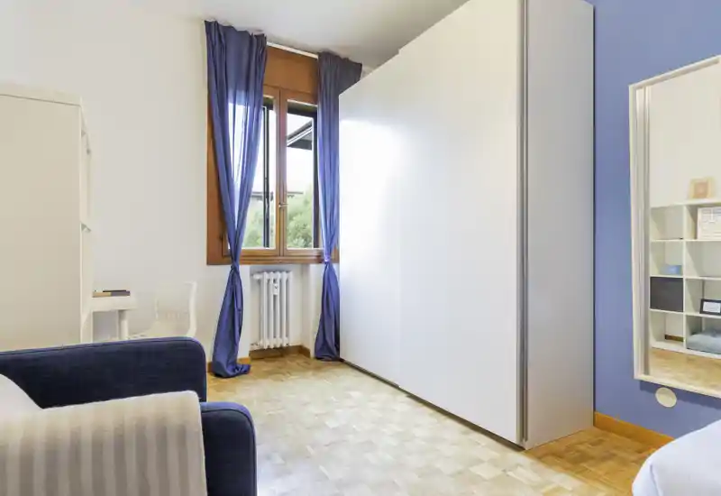 Bright, simply furnished bedroom with wooden parquet, large wardrobe and a window with blue curtains. Partial view of the bed and a cozy navy armchair create a welcoming, airy atmosphere.